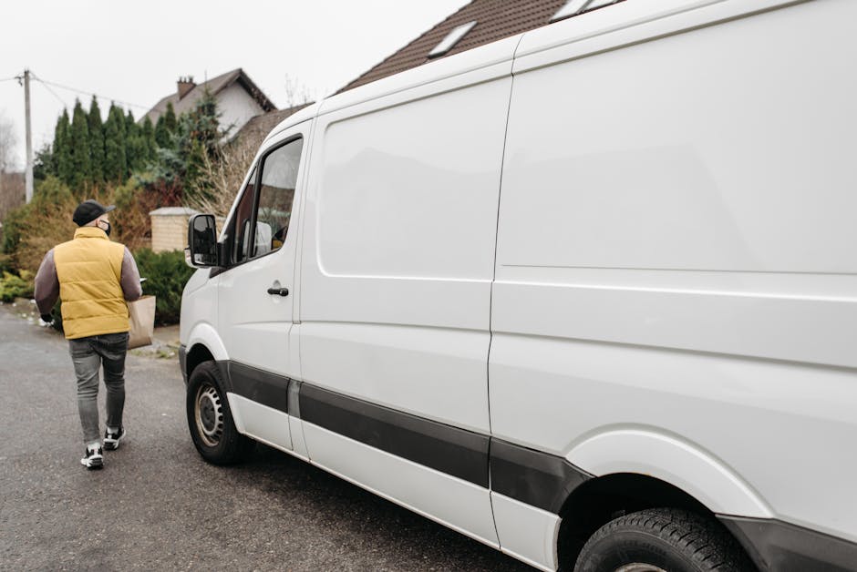 A man wearing a yellow padded vest, grey joggers, and a black cap is seen from behind walking towards a white commercial van parked on a residential street. The van, positioned parallel to the curb, is a large panel type with sliding side doors and black trim along the lower edge. The street surface is paved, and nearby are houses with pitched roofs, some with visible chimneys, along with greenery including trees and bushes. The man is carrying a cardboard box, likely part of a home relocation or furniture transport process involving detailed packing and moving activities. The van is ready for loading or unloading furniture, boxes, or appliances, supporting the logistics involved in house removals. This scene is indicative of a professional removal service like Man With a Van Shacklewell, focusing on the loading process in a typical residential environment.