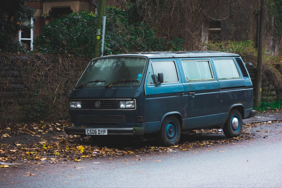 A dark blue vintage Volkswagen Type 2 van parked on a residential street curb, surrounded by fallen autumn leaves. The van has a boxy shape typical of classic camper vans, with a flat front, rectangular headlights, and a prominent VW emblem on the grille. The vehicle features a sliding side window with frosted glass, a small side mirror, and hubcaps matching the body colour, with rearview mirrors on each side. It is situated close to a pavement alongside a weathered wooden fence and overgrown shrubbery, indicating a peaceful neighbourhood setting. The van appears to be unused for some time, with slight rust visible on the bodywork. This vehicle is positioned in front of an area possibly used for loading or unloading, relevant to home relocation and furniture transport processes. Occasionally, Man With a Van Shacklewell handles moving services involving such vehicles, fitting within their scope of removals and packing logistics.