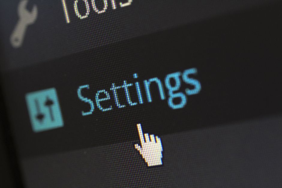 Close-up of a digital device screen displaying a blue-highlighted 'Settings' menu option, with a white pixelated hand cursor pointing at it. The background features part of the device interface, with a dark theme and other menu icons partially visible. This image relates to configuring or adjusting options in a technical or moving-related context, such as packing or logistics settings, and may be used in content about house removals or moving services provided by Man With a Van Shacklewell.