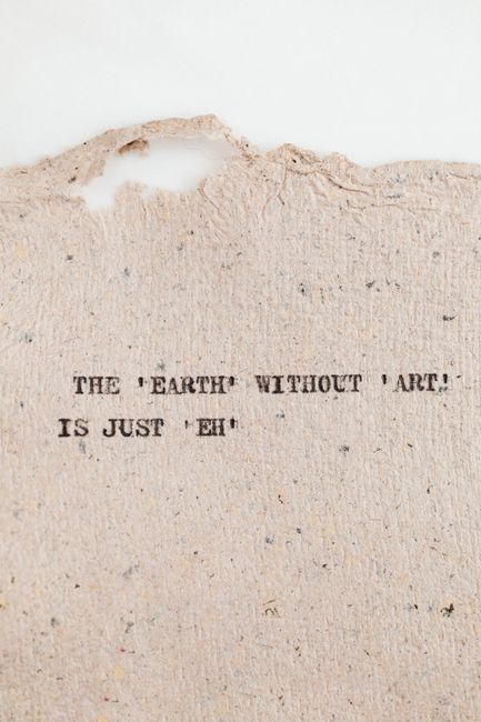 Close-up of a piece of textured, slightly torn paper with a quote printed in black ink that reads, 'The earth without art is just eh.' The paper shows a rough edge at the top with a small tear and some fibrous detail, suggesting it may be part of a handmade or recycled material. The lighting highlights the surface texture and subtle variations in the paper's color, which is off-white or cream. The quote is centered and the font is simple and typewriter-like, adding a contemplative tone. This image does not depict any objects related to house removals or moving services but may serve as a decorative or motivational element in a related context.