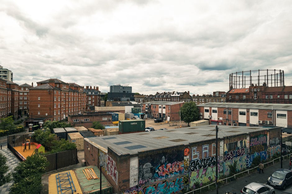 A wide view of an urban setting showing a mix of residential and commercial buildings under a cloudy sky. In the foreground, a low-rise building with a graffiti-covered roof is visible, surrounded by a fenced outdoor area that includes a small playhouse and a garden with trees and shrubs. Behind this, there are several more low-rise structures, some with flat roofs and others with slightly pitched roofs, featuring exterior walls in various shades of brick and concrete. To the right, a large tank or silo structure is visible on the rooftop of one of the buildings. In the middle ground, there are paved open spaces with parking areas containing cars, and shipping containers are stacked near some warehouses or storage units. Towards the background, multiple multi-storey residential buildings made of brick and concrete are seen, with windows and balconies, along with a tall, circular industrial tank or cooling tower. The overall scene appears to depict a typical urban neighbourhood, with ongoing or recent home relocation or building activity that Man With a Van Shacklewell offers as part of their removals services.