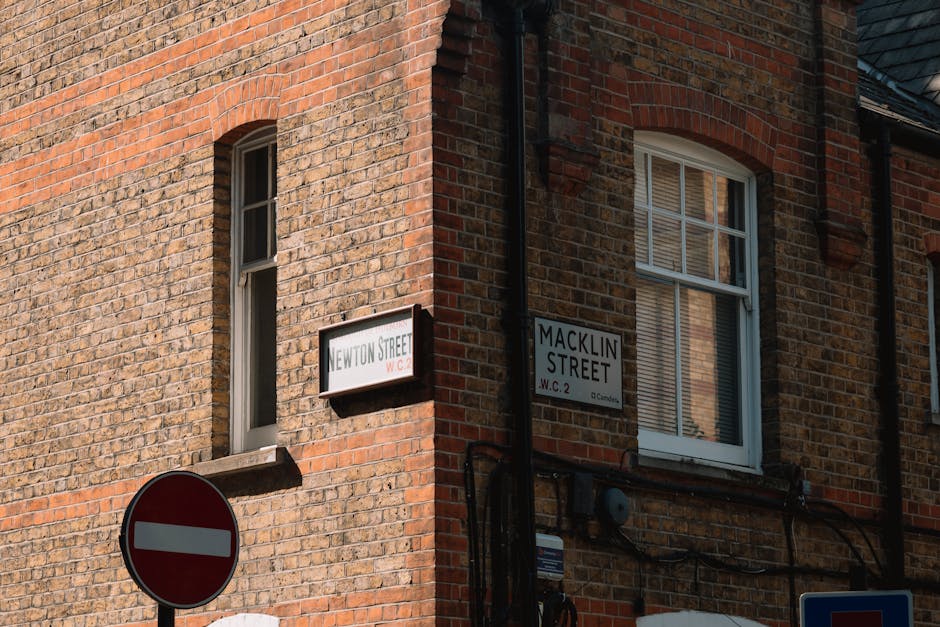Close-up view of the corner of a brick building at the intersection of Newton Street and Macklin Street in London, displaying two street signs attached to the brick wall. The Newton Street sign is positioned on the left side and the Macklin Street sign on the right, both in black letters on a white background. The building features a window with white framing and partially visible blinds, situated above a black drainpipe running down the corner. The brickwork consists of red and brown tones, with some weathering and mortar visible. In the lower left corner, part of a red and white circular 'no entry' road sign is visible, alongside cables and electrical meters attached to the wall. The scene is illuminated by natural daylight, highlighting the textures of the brick exterior and the street signs, suggesting an urban setting typical for house removals or relocation services like those provided by Man With a Van Shacklewell.
