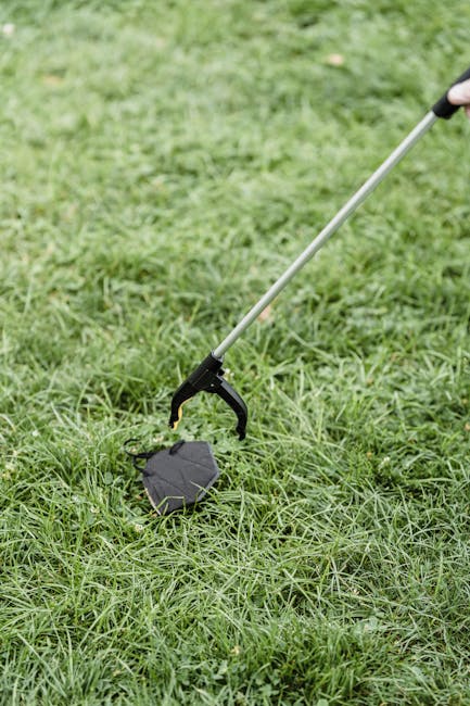 A person using a long-handled grabber tool with a black grip is picking up a dark grey, irregularly shaped object made of plastic or rubber from a grassy outdoor area. The grass is lush and green, with some taller patches, and the scene is outside a property, likely near a house or garden. In the background, a section of the lawn is visible, and the person's hand is partially visible, holding the grabber tool for assistance in lifting or disposing of the object. The scene depicts the process of garden clearance or waste disposal following a house move, with the use of equipment typical of removals and clearance services offered by Man With a Van Shacklewell.