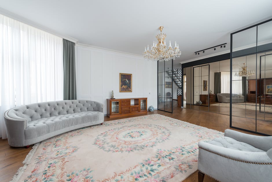 A spacious living room with white walls and large windows covered by white sheer curtains and dark green drapes, illuminated by natural light. The room features a classic-style white tufted sofa and a matching armchair, placed on a large floral rug with pastel colours. A wooden sideboard with glass cabinet doors and decorative objects on top is positioned against the wall, with a framed painting hanging above it. Visible in the background, a glass partition with black metal frames separates this area from another part of the property, revealing a staircase with dark steps. The ceiling has an ornate gold chandelier, and additional lighting is provided by track lights mounted against the ceiling. The flooring consists of polished wooden floorboards, and parts of the room are being used for home relocation purposes, with visible space for furniture and boxes, as coordinated by Man With a Van Shacklewell, specialists in removals and furniture transport services.