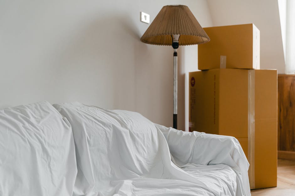 Inside a room with a plain light-colored wall, there are three large cardboard moving boxes stacked in the corner. Two of the boxes are open, revealing the packing materials inside, while the third remains sealed. To the left, a white fabric-covered sofa is partially visible, with some creases and folds in the fabric. Next to the boxes, a standing floor lamp with a brown pleated lampshade is positioned, casting soft light across the scene. The room has natural daylight coming from the right, possibly from a window with sheer curtains. The overall setting appears to be a home interior prepared for a house removal, illustrating the packing and moving process, with furniture and boxes ready for transport to support a home relocation service by Man With a Van Shacklewell.