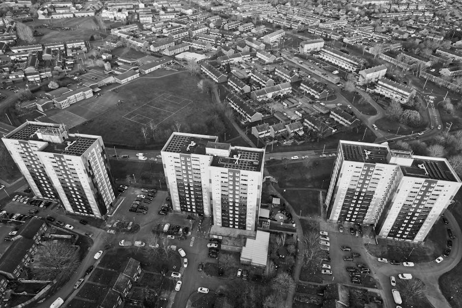 Black and white aerial photograph of a residential area showing three large, modern multi-storey apartment buildings in the foreground, each with multiple balconies and flat roofs, surrounded by parking spaces filled with various cars. Behind the buildings, there is a large open green space with a sports field marked with lines, bordered by trees and a mix of houses and smaller buildings arranged along streets. The scene captures a suburban environment during daytime, with overcast lighting creating soft shadows, depicting the typical setting for house removals and furniture transport as part of home relocation services offered by Man With a Van Shacklewell.