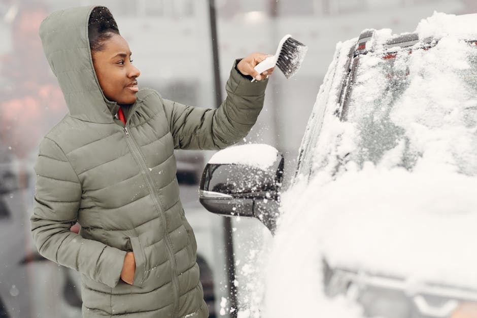 A person wearing a grey hooded puffer jacket is standing outside in snowy weather, using a brush to clear snow from the side mirror and windshield of a black vehicle parked on a pavement. The vehicle’s surface is covered with snow, and the person is holding the brush with one hand while the other hand is in their pocket. The background shows a blurry urban environment with other parked cars and buildings. This scene depicts winter weather conditions during a home relocation process, where careful clearing of vehicles is part of the moving logistics. Man With a Van Shacklewell offers removals services that include vehicle transportation and packing during winter months, ensuring safe and efficient home or furniture transport.