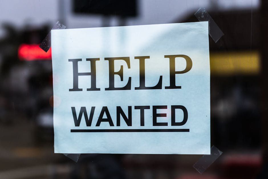 A white sheet of paper taped to a glass window with four pieces of black tape at each corner. The paper displays the message 'HELP WANTED' in bold, uppercase letters, with 'HELP' printed in black and 