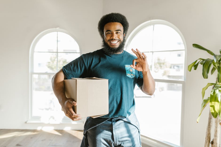 A smiling man with a natural Afro hairstyle and a full beard stands inside a bright, minimally furnished room with large arched windows, holding a cardboard box in his left hand. He is wearing a teal t-shirt and casual jeans, and appears to be part of a house removal or furniture transport process. To his right, a green plant adds a touch of decor, while in the background, sunlight streams through the windows, illuminating the space. The man is making an OK gesture with his right hand, indicating a positive or successful move. The scene reflects a professional home relocation or packing and moving service environment, with natural lighting highlighting the interior surroundings and the movement process involved in loading or unpacking appliances and boxes as part of a typical property transfer handled by Man With a Van Shacklewell.