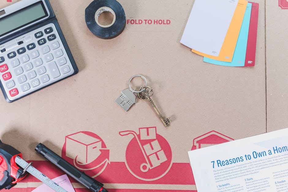 A top-down view of a cardboard moving box part of a house relocation process, featuring various items such as a black calculator with white and red buttons, a roll of black adhesive tape, a set of house keys with a silver keyring, a black marker pen, a folded tape measure with a pink section, and a printed document titled '7 Reasons to Own a Home' with blue text. In the background, there are colorful paper file folders in white, orange, blue, and pink, along with a partially visible white sheet of paper. The cardboard surface displays red recycling and moving icons, including a box, a hand truck, and a recycling symbol, highlighting logistical elements involved in furniture transport and packing during a home removal. The scene involves items associated with packing, organizing, and preparing belongings for moving, which aligns with services offered by Man With a Van Shacklewell.
