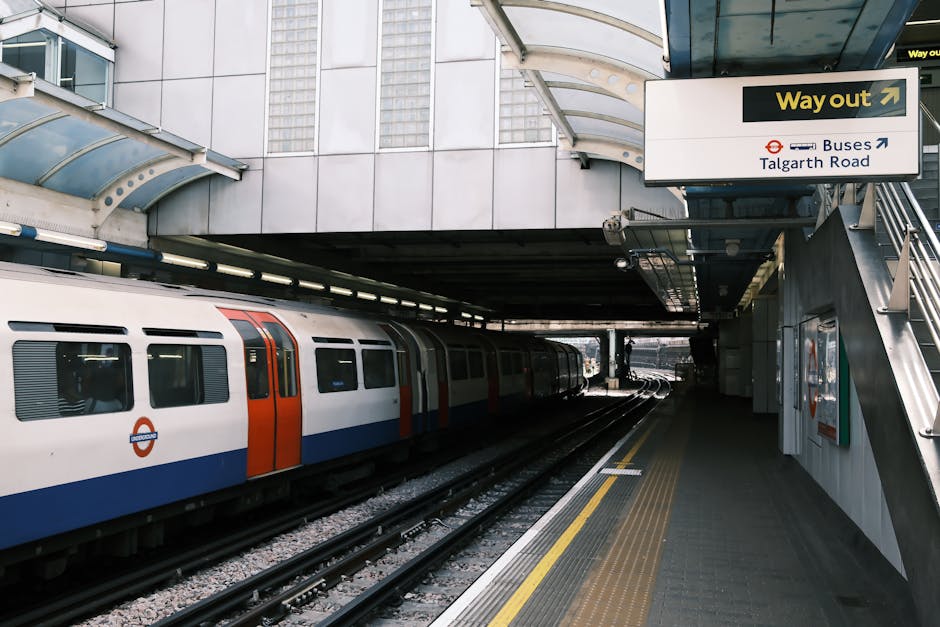 Exterior view of a London underground station platform adjacent to railway tracks, with a modern electric train featuring a white body, orange and blue markings, and station signage visible. The platform has a yellow safety line along the edge and a tactile paving strip for visually impaired passengers, while overhead signage indicates directions to buses and Talgarth Road. The station's structural elements include a curved glass and metal canopy, stairs leading up to an exit, and a sign that reads 'Way out.' The scene is illuminated by natural daylight, and the environment reflects a typical setting for house removals and transport logistics, illustrating the importance of planning travel routes in conjunction with moving services provided by companies like Man With a Van Shacklewell.