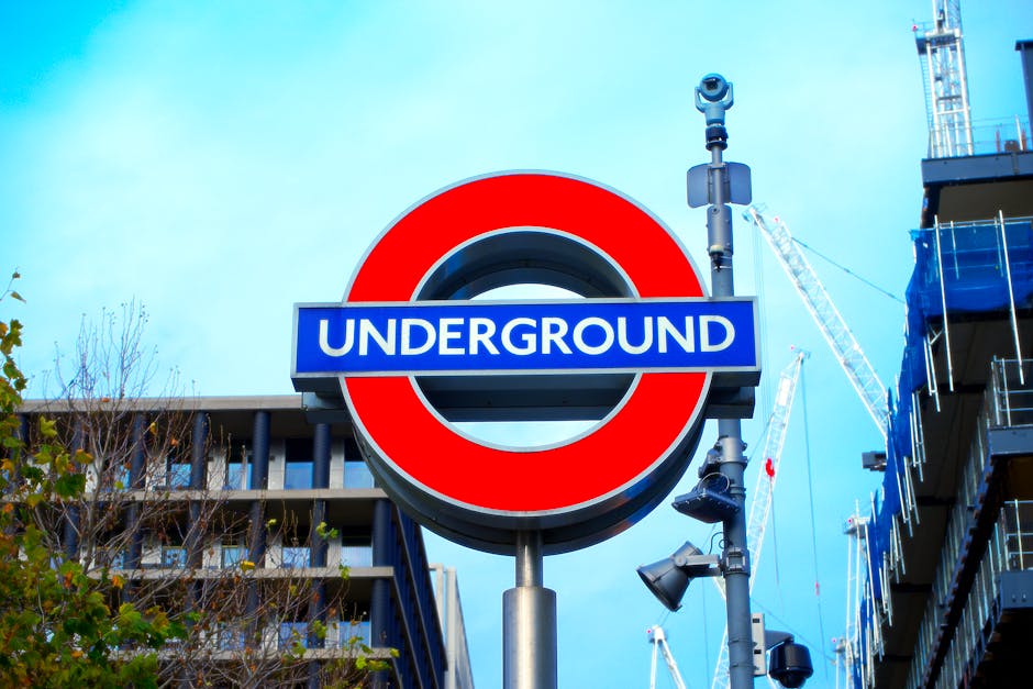 A large London Underground roundel sign with a prominent red circle and blue bar displaying the word 'UNDERGROUND' in white text, mounted on a metal pole against a backdrop of modern multi-storey buildings with glass balconies on the right and scaffolding with cranes on the left, indicative of ongoing construction. The sky is clear with a few clouds, and nearby streetlights and cameras are attached to the pole. The scene is set in an urban environment, likely near a station or transport hub, with foliage visible at the lower left corner. This image exemplifies urban infrastructure, relevant to relocation and moving logistics in central London, with contemporary building developments and transport signage supporting home relocation and furniture transport services offered by Man With a Van Shacklewell.