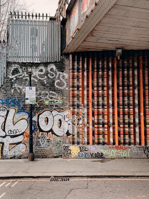 Exterior view of a London underground station platform adjacent to railway tracks, with a modern electric train featuring a white body, orange and blue markings, and station signage visible. The platform has a yellow safety line along the edge and a tactile paving strip for visually impaired passengers, while overhead signage indicates directions to buses and Talgarth Road. The station's structural elements include a curved glass and metal canopy, stairs leading up to an exit, and a sign that reads 'Way out.' The scene is illuminated by natural daylight, and the environment reflects a typical setting for house removals and transport logistics, illustrating the importance of planning travel routes in conjunction with moving services provided by companies like Man With a Van Shacklewell.