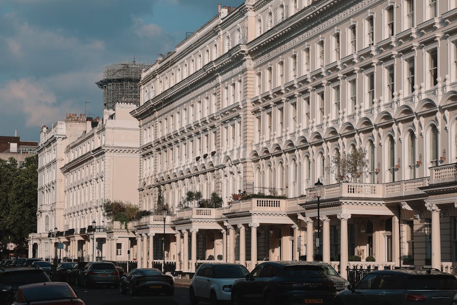 The image shows a row of elegant Victorian terraced houses with white facades, ornate architectural details, and large windows, situated on Shacklewell Road. In front of the buildings, there is a street with several parked cars on the pavement. Visible within the scene is an open doorway leading into the interior of one of the houses, where moving boxes made of cardboard and plastic wrapping can be seen inside. The boxes are stacked near the entrance, some covered with blankets or protective materials, indicating packing for a home relocation. A moving trolley or dolly is partially visible, positioned nearby, suggesting staff from Man With a Van Shacklewell are involved in furniture transport or loading activities. The scene is well-lit with natural daylight, alongside street lamps on the sidewalk, emphasizing a typical urban environment prepared for a packing and moving process involving the loading of household items into the van. A construction scaffold is visible on a neighboring building, adding to the street’s characteristic architecture and ongoing developments.
