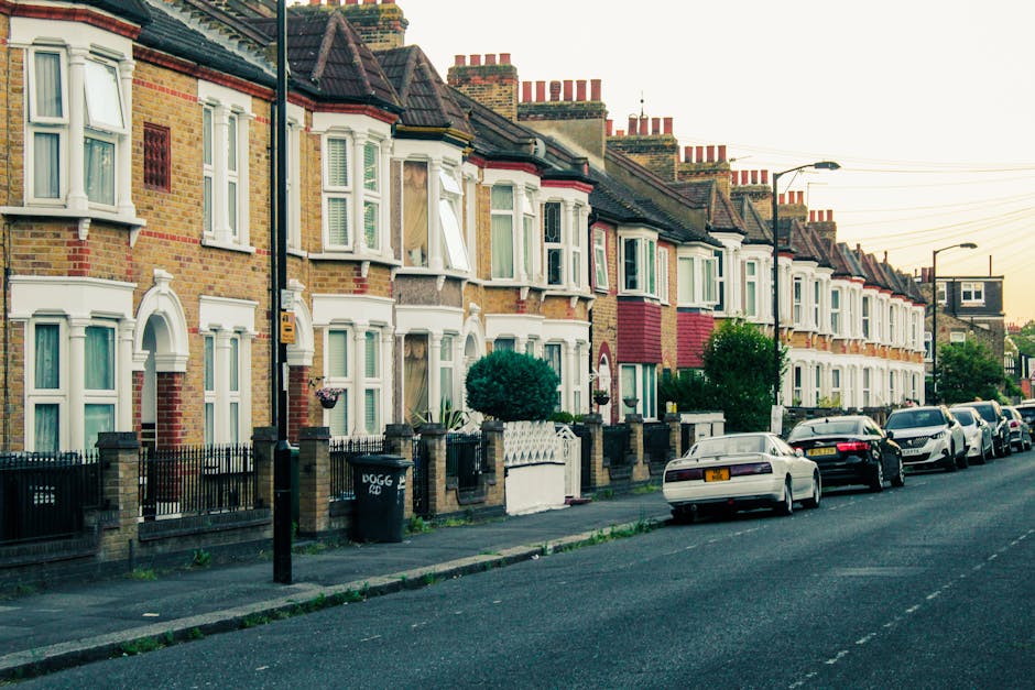 A row of traditional Victorian terraced houses with brick facades, white-framed bay windows, and tiled roofs, lining a residential street during daylight. Several cars are parked along the curbside, including hatchbacks and sedans, with some partially visible in the foreground. The sidewalk features small front gardens separated by low brick walls and black metal railings, and a black wheelie bin is positioned near one entrance. A lamppost and a street sign are also visible, with overhead power lines stretching across the scene. The street appears quiet and well-maintained, with a subtle glow of natural morning or evening light. This setting is typical of a London suburb suitable for home relocation and furniture transport, as part of packing and moving services provided by Man With a Van Shacklewell, supporting efficient house removals and relocation logistics.