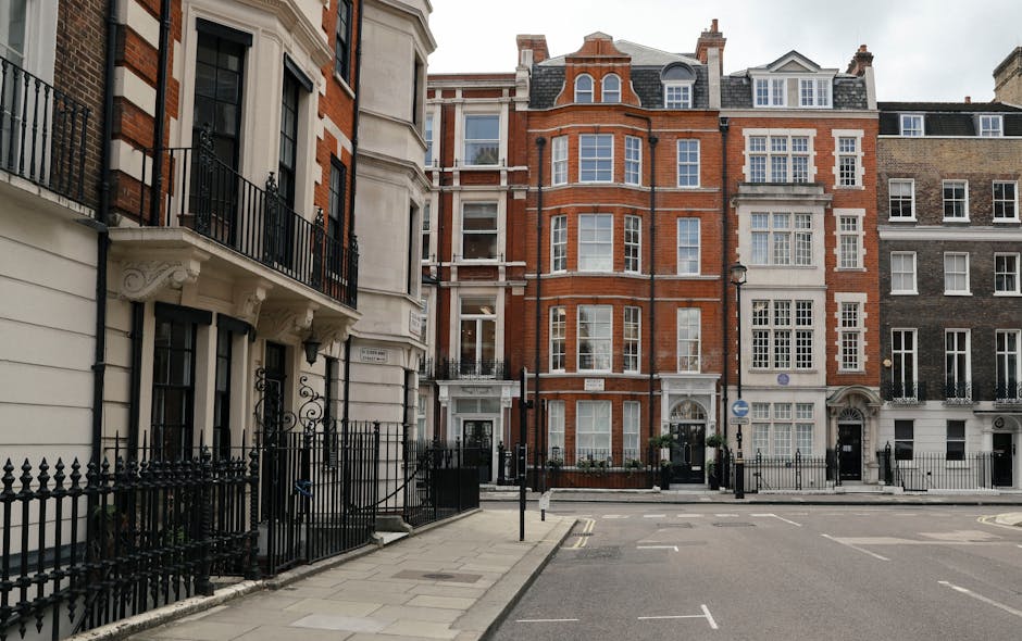 The image shows a row of elegant Victorian terraced houses with white facades, ornate architectural details, and large windows, situated on Shacklewell Road. In front of the buildings, there is a street with several parked cars on the pavement. Visible within the scene is an open doorway leading into the interior of one of the houses, where moving boxes made of cardboard and plastic wrapping can be seen inside. The boxes are stacked near the entrance, some covered with blankets or protective materials, indicating packing for a home relocation. A moving trolley or dolly is partially visible, positioned nearby, suggesting staff from Man With a Van Shacklewell are involved in furniture transport or loading activities. The scene is well-lit with natural daylight, alongside street lamps on the sidewalk, emphasizing a typical urban environment prepared for a packing and moving process involving the loading of household items into the van. A construction scaffold is visible on a neighboring building, adding to the street’s characteristic architecture and ongoing developments.