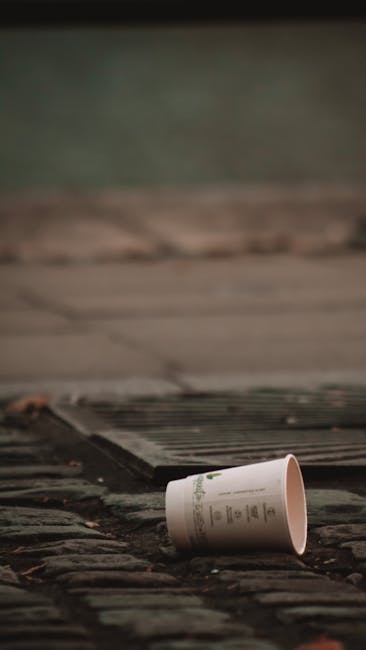 A disposable paper coffee cup with a white sleeve lies on its side on a wet wooden doorstep, near a slightly raised rubber mat at the entrance of a property. The doorstep is made of dark wood or stone, and the background shows a blurred view of the exterior wall of the house with a muted green and beige color scheme. The surrounding area appears damp, suggesting recent rain. This scene depicts an outdoor setting during a home relocation or moving process, where securing and clearing the entrance is part of the logistics managed by Man With a Van Shacklewell. The image emphasizes the importance of maintaining a tidy entrance when arranging removals and furniture transport, with attention to packing and loading procedures to ensure smooth access for moving vehicles and personnel.