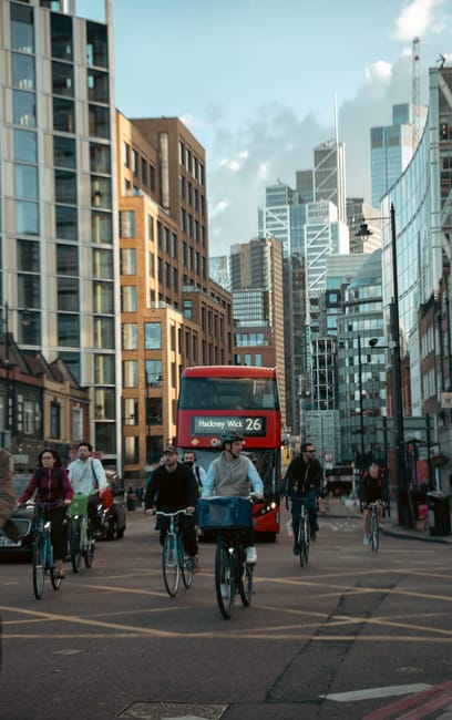 Image showing a busy urban street scene in Hackney with several people riding bicycles, some carrying bags on their bikes, and others walking. A red double-decker bus with route information for Hackney Wick 26 is approaching in the background amid tall modern office buildings and older brick structures. The scene is illuminated by daylight with partly cloudy skies, and pedestrians and cyclists are traveling along the city street, which features designated bike lanes and sidewalks. This environment reflects a typical city area where home relocation and furniture transport services by Man With a Van Shacklewell could be coordinated during city-based moving or packing and moving activities involving delivery vehicles and urban logistics.
