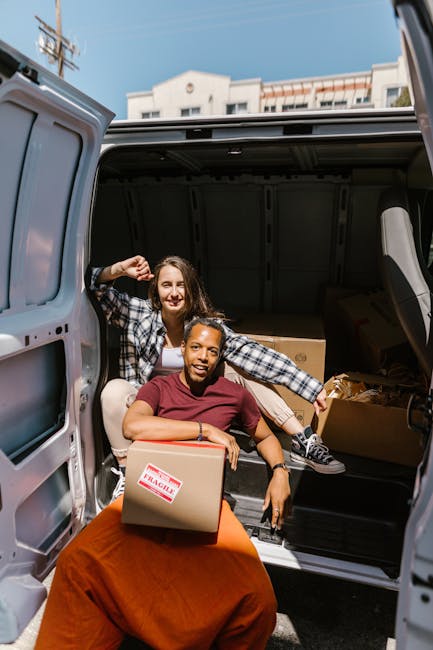 Two young women are loading cardboard boxes into the open rear cargo area of a grey van parked outside a building. One woman with long, dark hair is seated at the edge of the van's loading area, holding a medium-sized box with a 'fragile' sticker, while the other woman with shoulder-length hair, wearing a checkered shirt, is standing inside the van, smiling and holding onto a box. The interior of the van shows additional boxes and packing materials, such as plastic wrap and cardboard. The scene is set outdoors in daylight, with a clear sky and urban building rooftops visible in the background. The image illustrates the process of home relocation or furniture transport, coordinated by [COMPANY_NAME], a professional removals service, emphasizing packing and loading activities typical of move logistics.