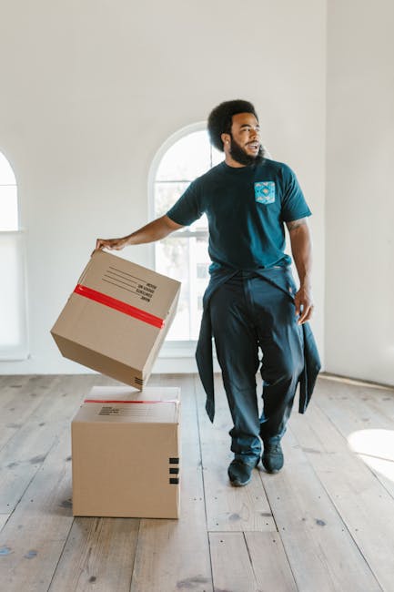 A man with an afro hairstyle and beard, dressed in a navy blue t-shirt and dark trousers, is inside a bright, minimally furnished room with white walls and large arched window. He is holding a cardboard box with red tape around it, preparing to place or lift it onto another similar box on the wooden floor. The room appears to be part of a home during a furniture transport or packing and moving process, with natural light coming through the window, highlighting the clean and spacious environment. This scene reflects the activities involved in home relocation, supported by professional removals services such as Man With a Van Shacklewell, demonstrating the careful handling of boxes during the loading process.